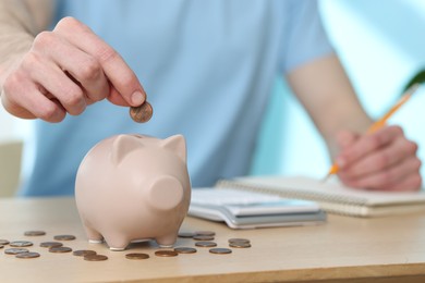 Photo of Financial savings. Man putting coin into piggy bank while writing down notes at wooden table, closeup