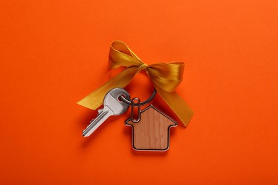 Key with trinket in shape of house and yellow bow on orange background, top view. Housewarming party