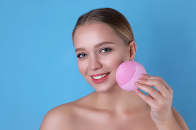 Young woman washing face with cleansing brush on light blue background. Cosmetic product