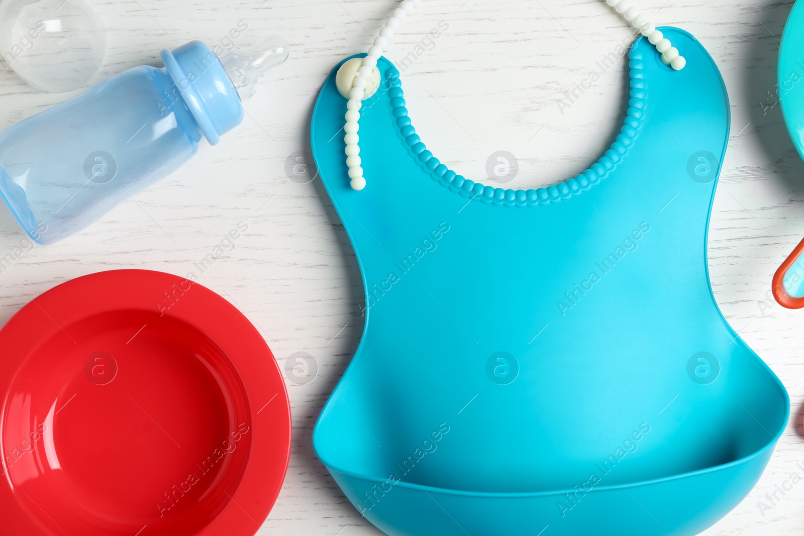 Photo of Set of plastic dishware and silicone bib on white wooden table, flat lay. Serving baby food