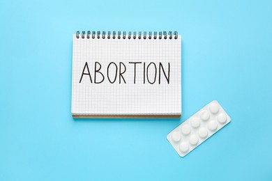 Photo of Notebook with word Abortion and pills on light blue background, flat lay