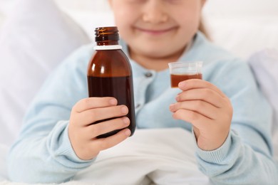 Photo of Closeup view of girl holding bottle and measuring cup with cough syrup in bed
