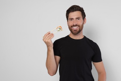 Photo of Happy man holding sushi roll with chopsticks on light grey background. Space for text