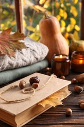 Photo of Book with autumn leaf as bookmark, scented candles and warm sweaters on wooden table near window