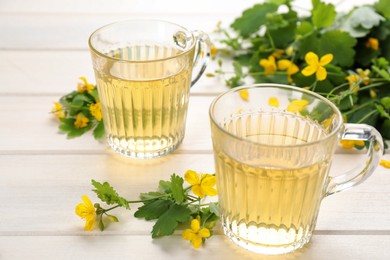 Photo of Glass cups of aromatic celandine tea and flowers on white wooden table