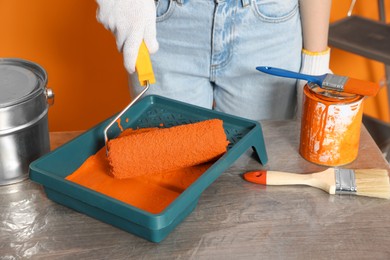 Photo of Woman taking orange paint with roller from tray at table, closeup