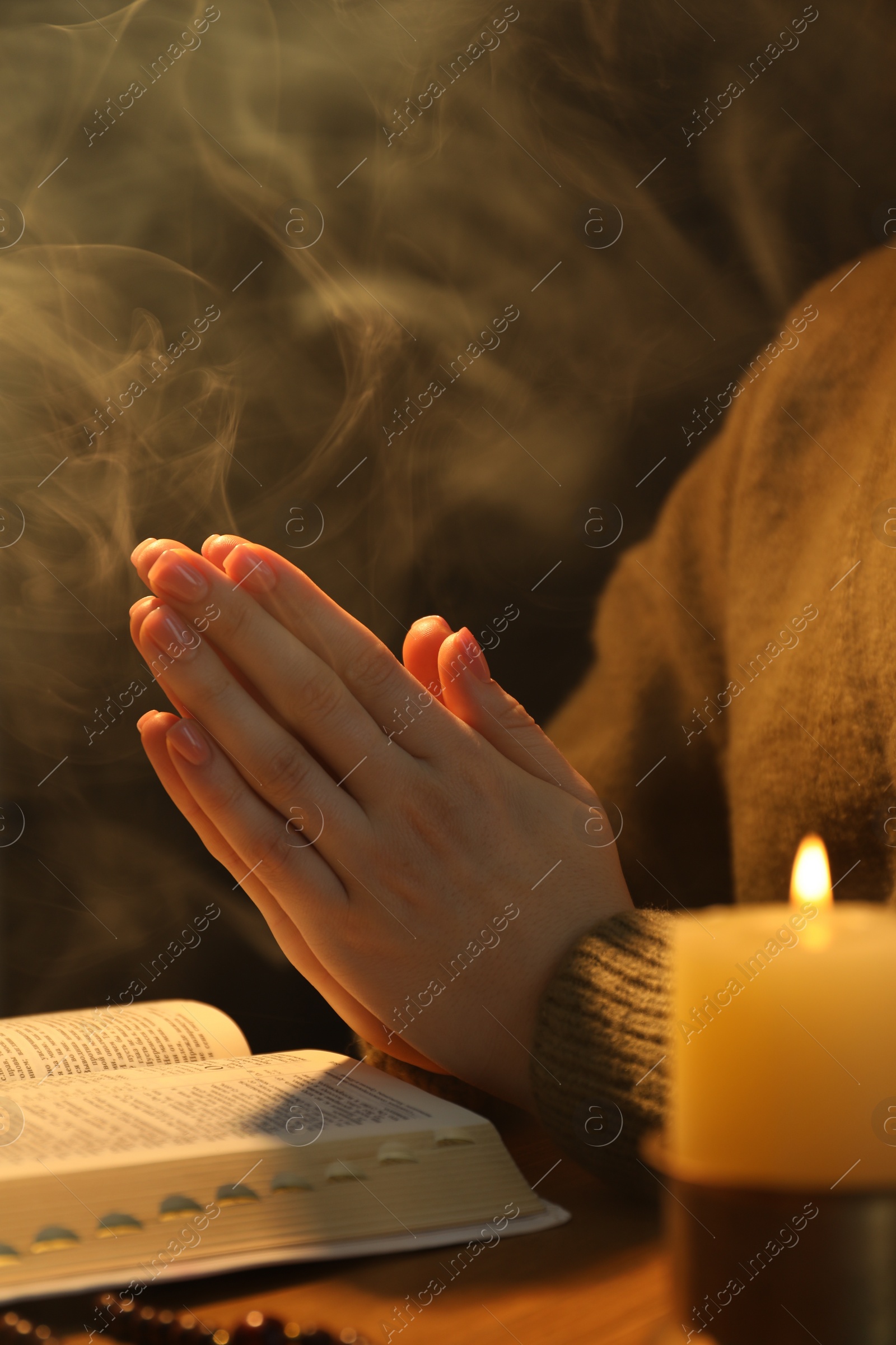 Photo of Woman praying at table with burning candle and Bible, closeup