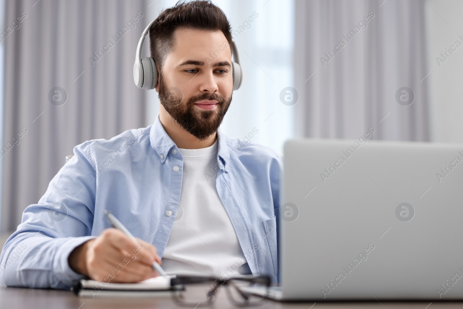 Photo of Young man in headphones watching webinar at table in room