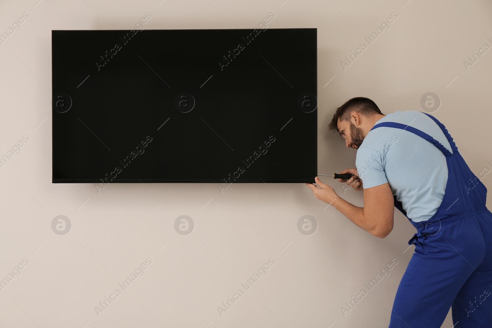 Photo of Professional technician with screwdriver installing modern flat screen TV on wall indoors