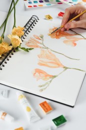 Woman painting freesias in sketchbook at white table, closeup
