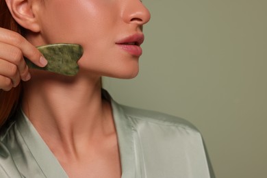 Photo of Young woman massaging her face with jade gua sha tool on green background, closeup. Space for text