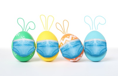 Image of COVID-19 pandemic. Colorful Easter eggs with cute bunny ears in protective masks on white background