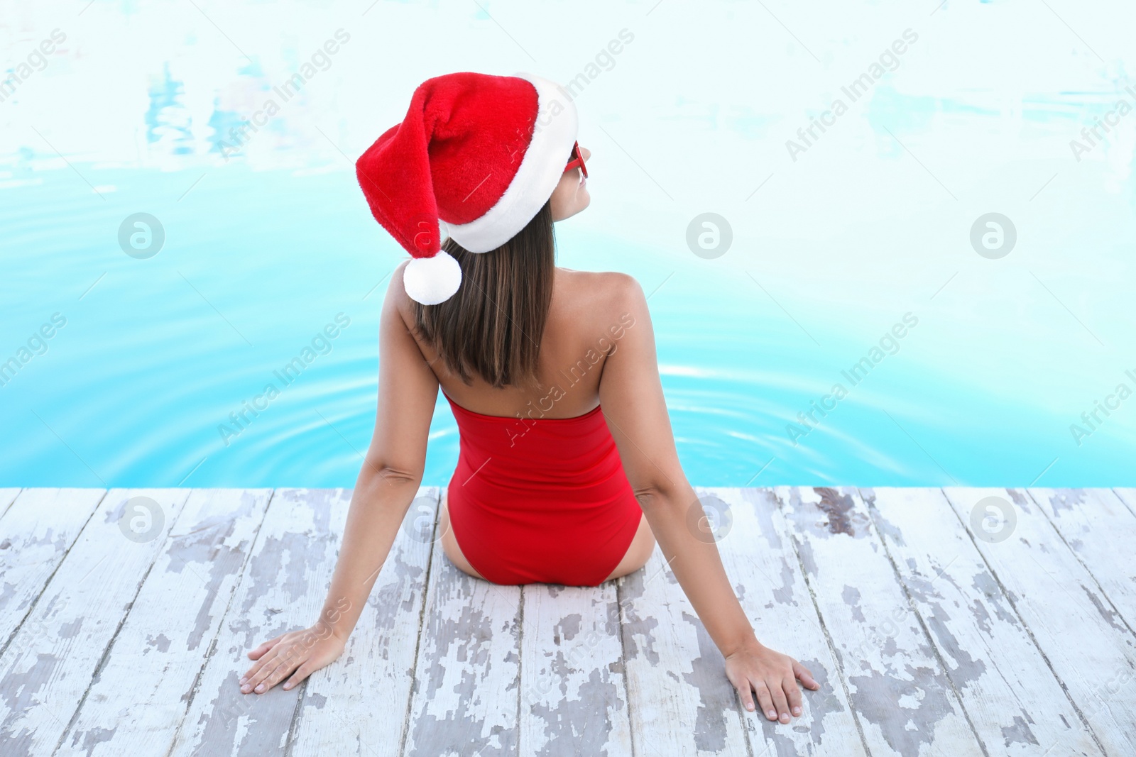 Photo of Young woman wearing Santa Claus hat near swimming pool, back view. Christmas vacation