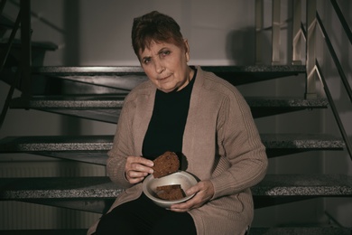 Poor senior woman with bread sitting on stairs indoors