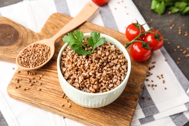 Board with bowl of buckwheat porridge, spoon and tomatoes on table