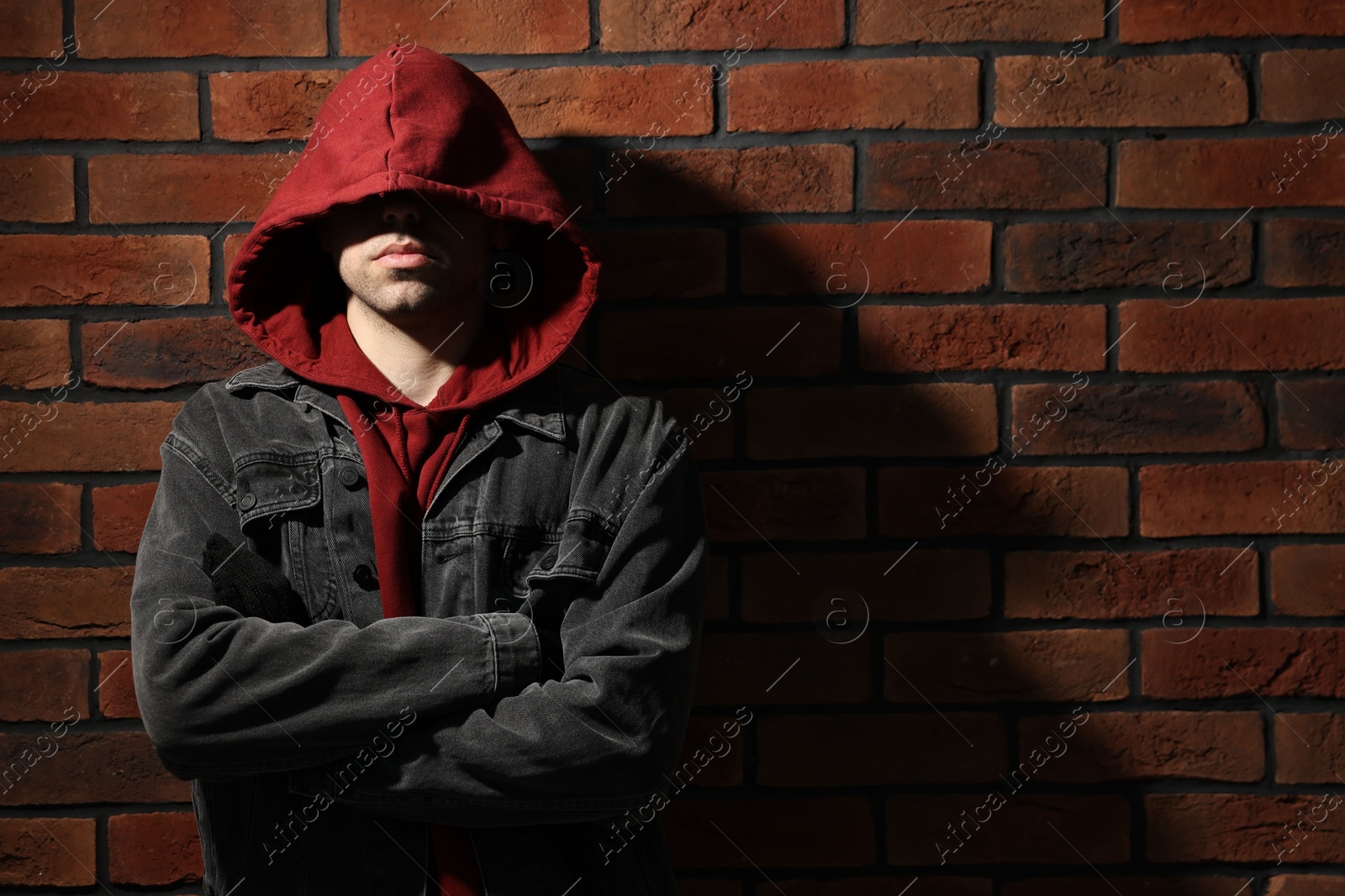 Photo of Thief in hoodie with crossed arms against red brick wall. Space for text