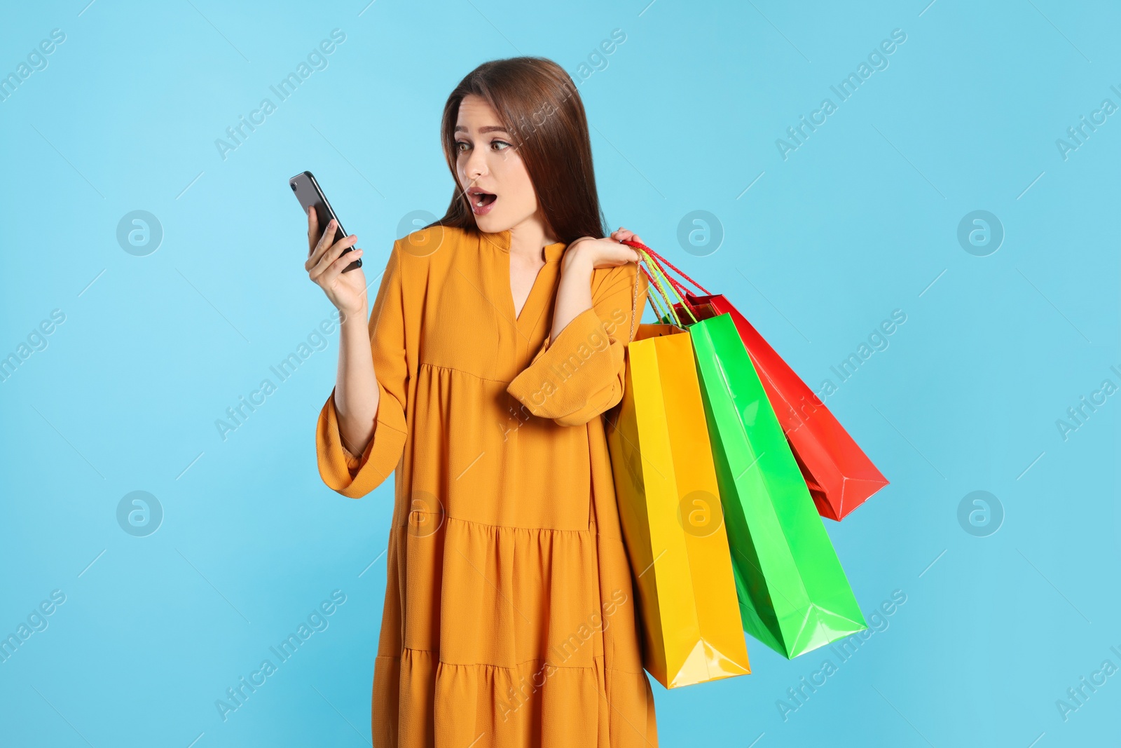 Photo of Surprised young woman with shopping bags and smartphone on light blue background. Big sale