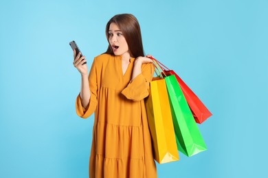 Surprised young woman with shopping bags and smartphone on light blue background. Big sale