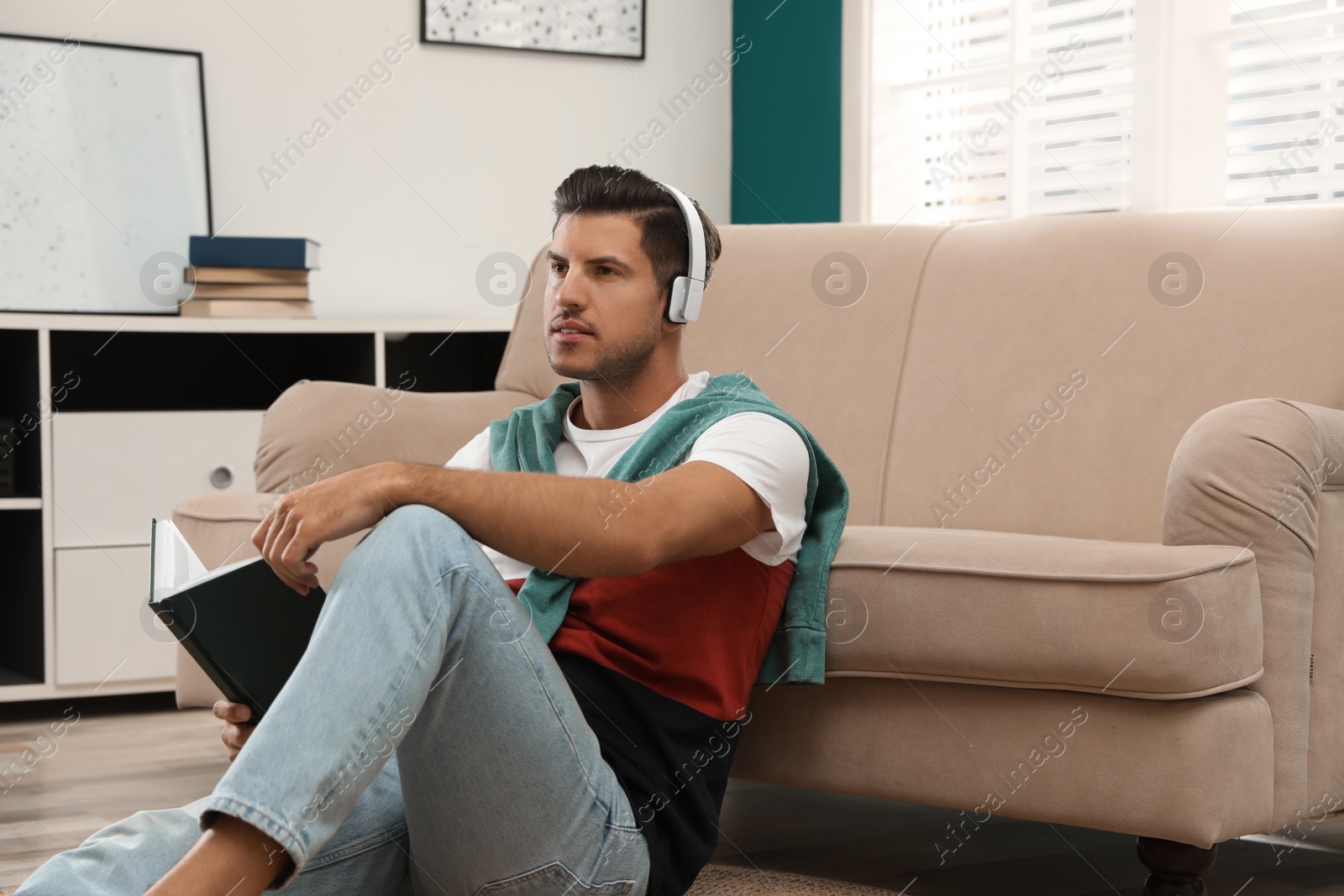 Photo of Man with headphones and book near sofa indoors. Audiobook concept
