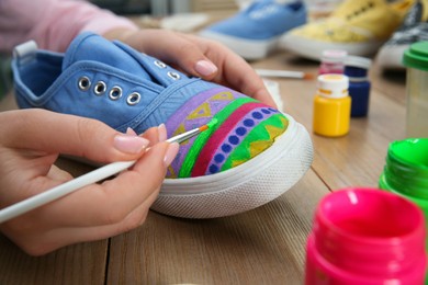 Photo of Woman painting on sneaker at wooden table, closeup. Customized shoes