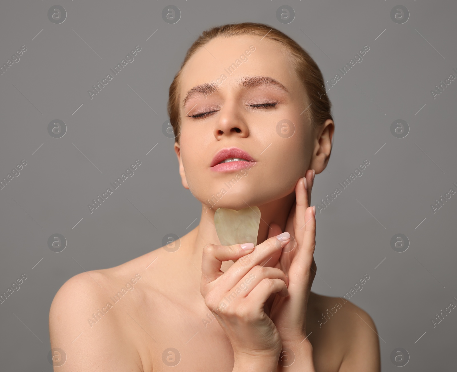 Photo of Beautiful young woman doing facial massage with gua sha tool on grey background