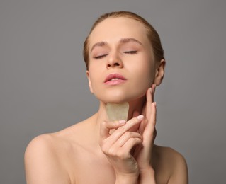 Beautiful young woman doing facial massage with gua sha tool on grey background