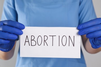 Photo of Doctor holding paper card with word ABORTION on light grey background, closeup