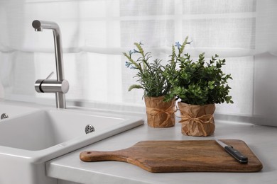 Photo of Artificial potted herbs, board and knife on white marble countertop in kitchen. Home decor