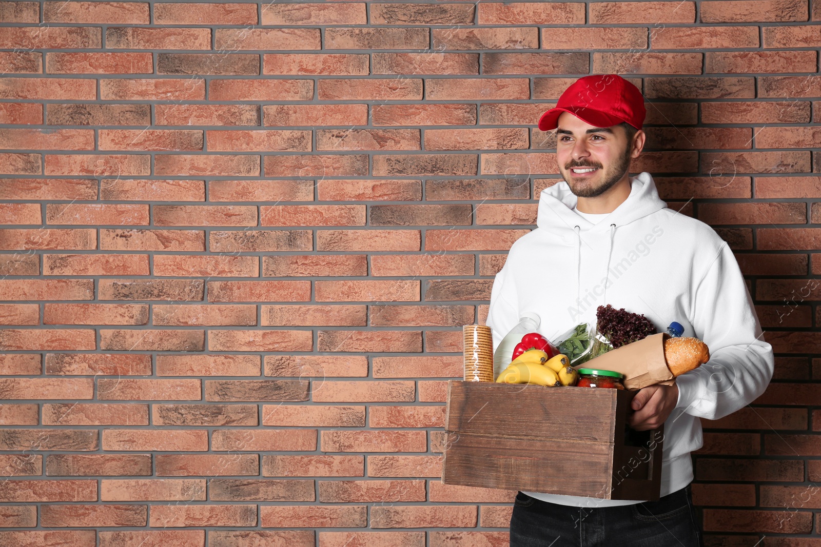 Photo of Food delivery courier holding wooden crate with products near brick wall. Space for text