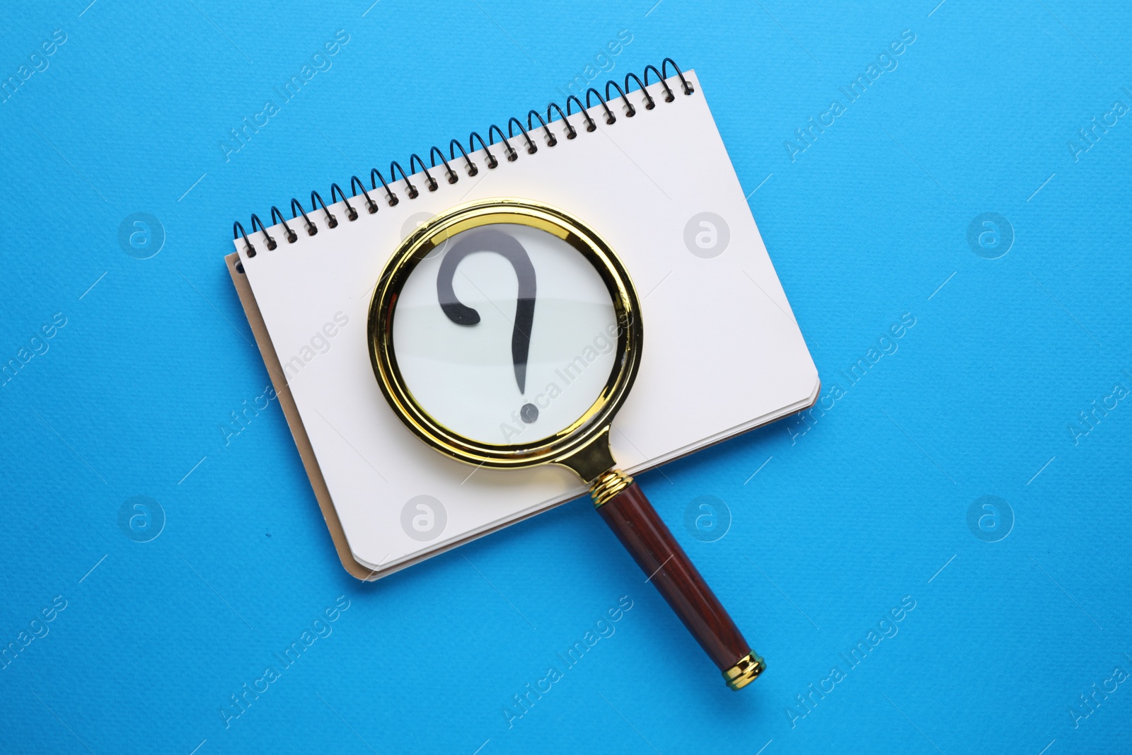 Photo of Magnifying glass over notebook with question mark on light blue background, top view