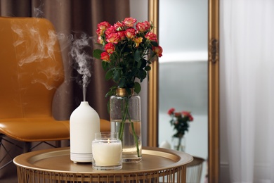 Photo of Aroma oil diffuser, burning candle and beautiful roses on table indoors, space for text. Interior elements