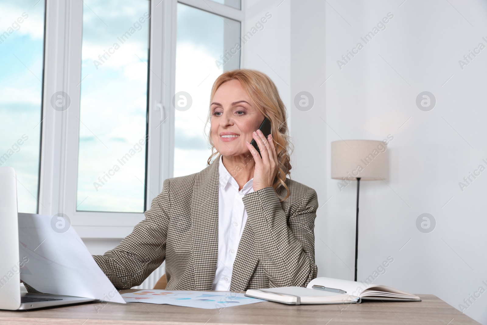 Photo of Lady boss with papers talking on smartphone at desk in office. Successful businesswoman