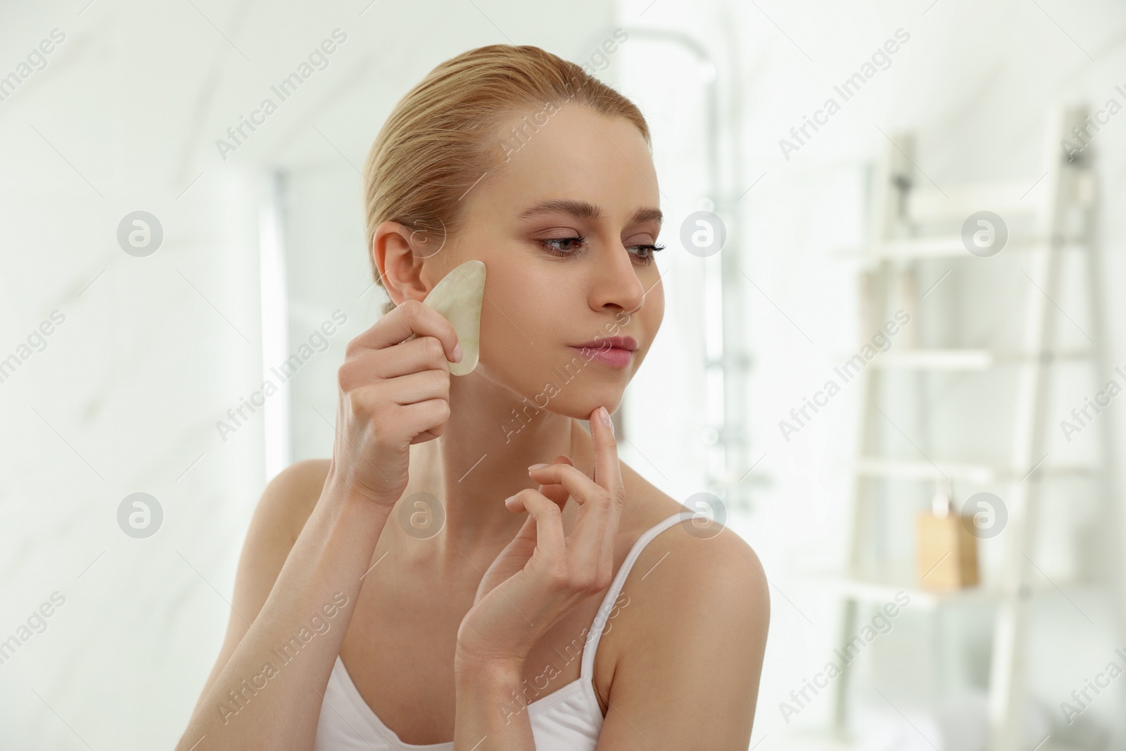 Photo of Beautiful young woman doing facial massage with gua sha tool in bathroom