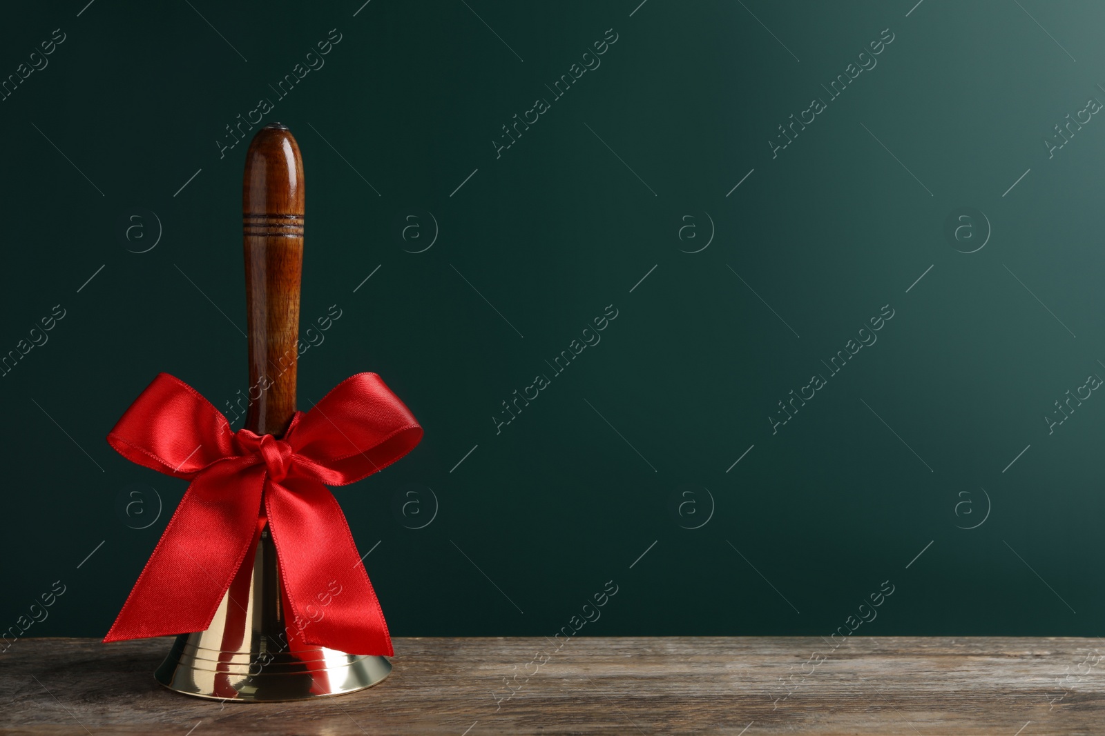 Photo of Golden bell with red bow on wooden table near green chalkboard, space for text. School days