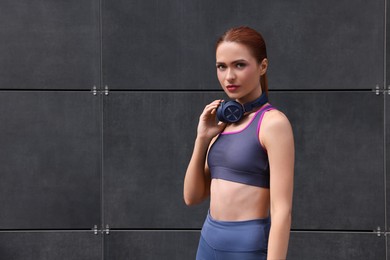 Photo of Beautiful woman in stylish gym clothes with headphones near dark grey wall on street, space for text