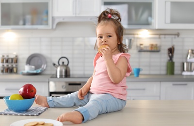 Photo of Adorable little baby girl eating tasty cookie on table in kitchen