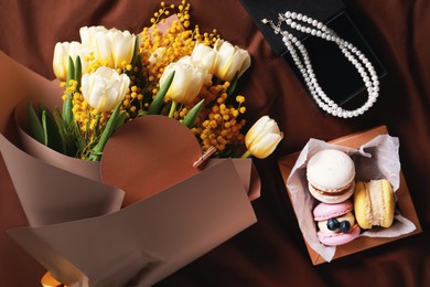 Photo of Bouquet of beautiful spring flowers, macarons and necklace on brown fabric, flat lay
