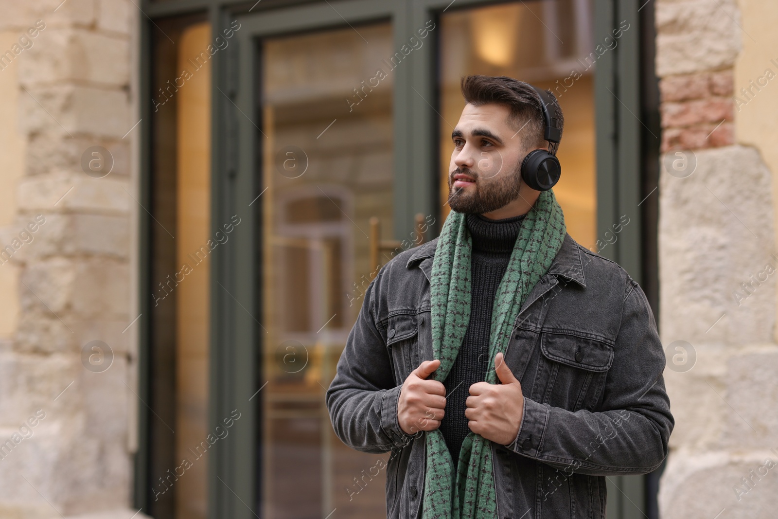 Photo of Handsome man in warm scarf and headphones near building outdoors. Space for text