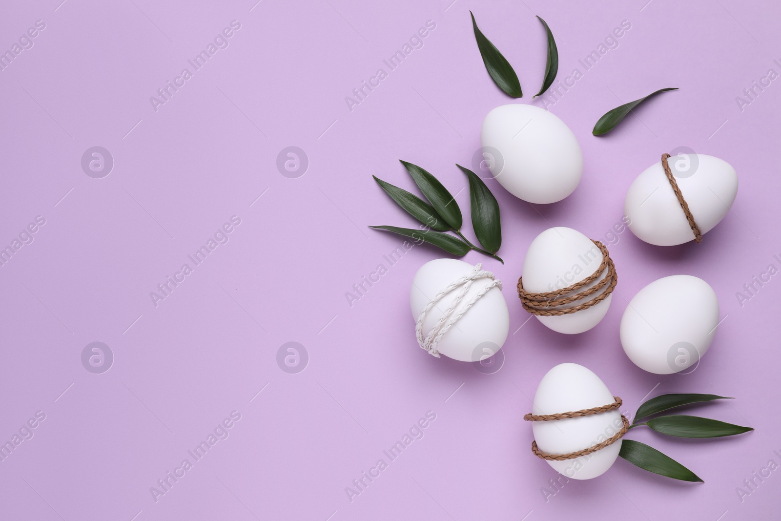 Photo of Beautifully decorated Easter eggs and green leaves on lilac background, flat lay. Space for text