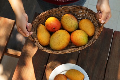 Woman with wicker basket of tasty mangoes at wooden table, top view