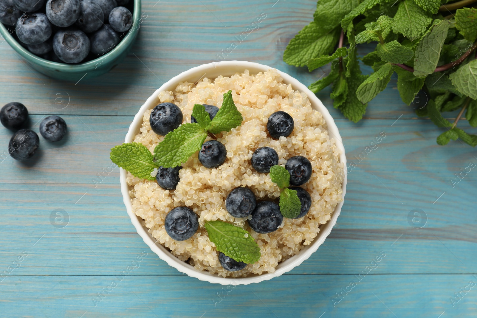 Photo of Tasty quinoa porridge with blueberries and mint in bowl on light blue wooden table, flat lay