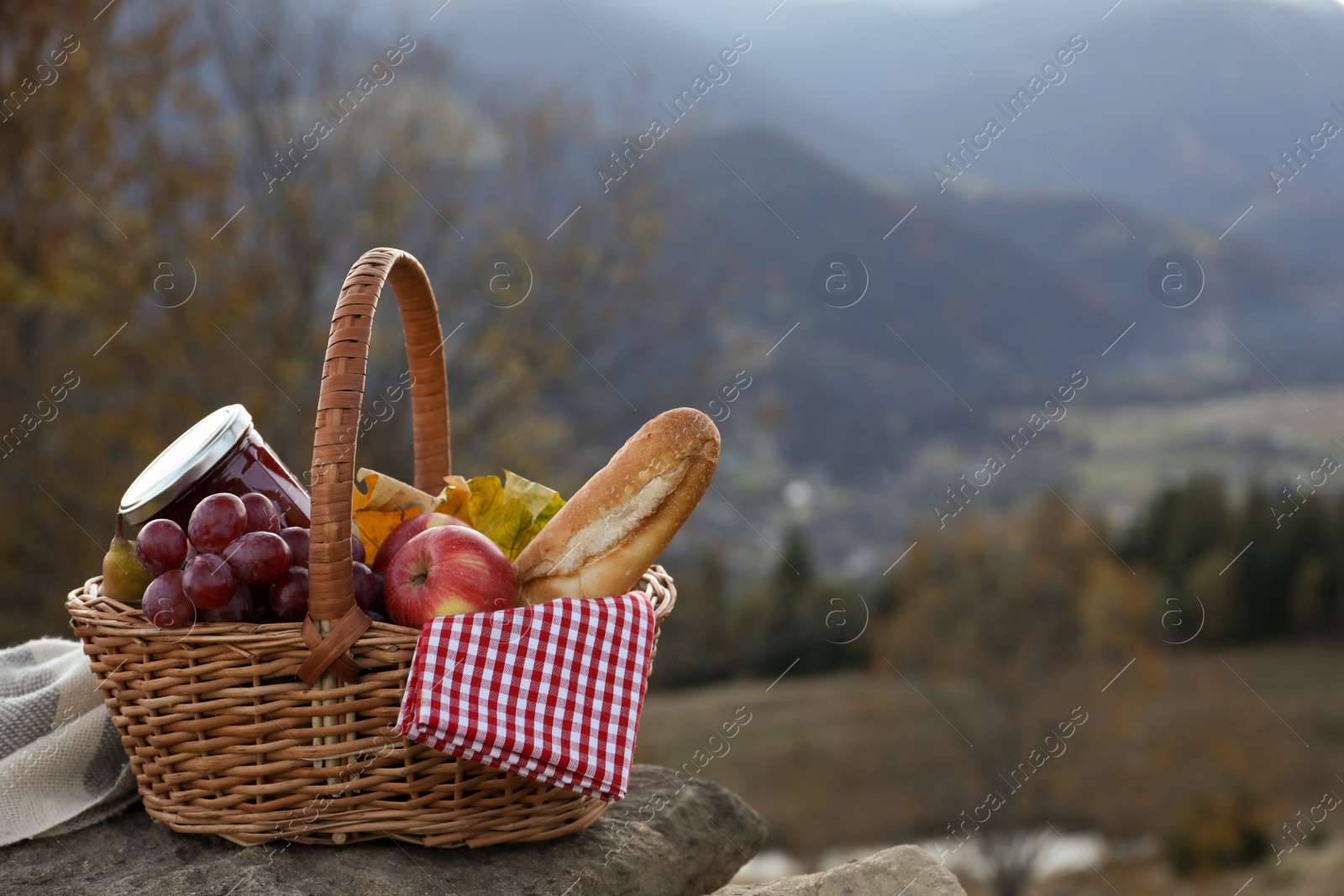 Photo of Wicker picnic basket with different products on rock in mountains. Space for text