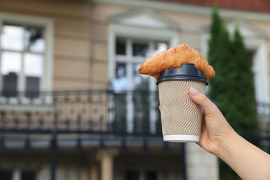 Woman holding croissant and paper cup in hand outdoors, closeup. Space for text