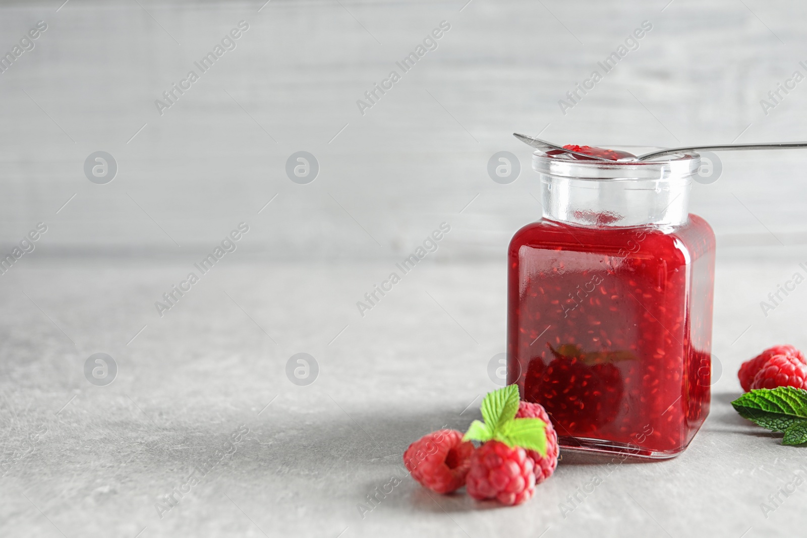 Photo of Glass jar and spoon of sweet jam with ripe raspberries on table. Space for text