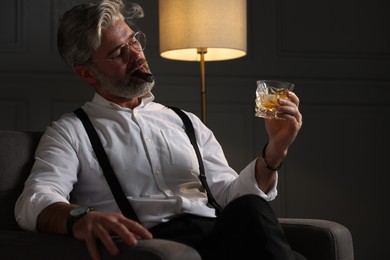Photo of Bearded man with glass of whiskey smoking cigar in armchair indoors