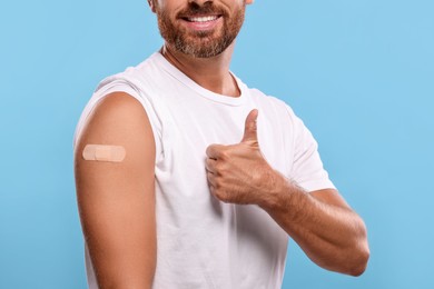 Photo of Man with sticking plaster on arm after vaccination showing thumbs up against light blue background, closeup