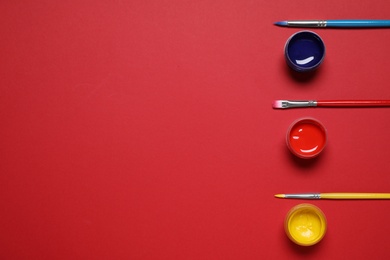 Brushes and jars with paint on red background, flat lay. Space for text