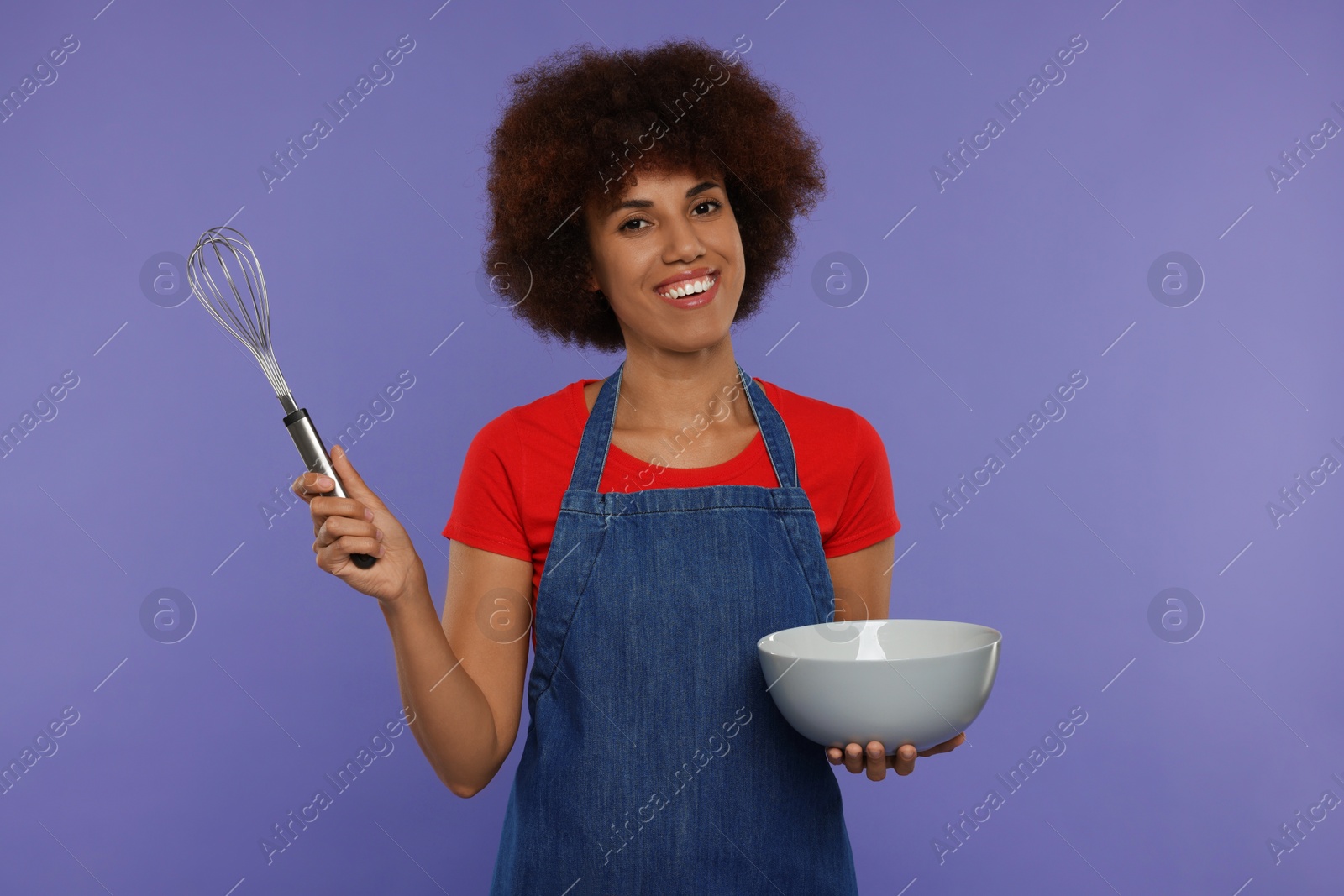 Photo of Happy young woman in apron holding whisk and bowl on purple background