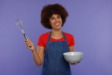 Happy young woman in apron holding whisk and bowl on purple background