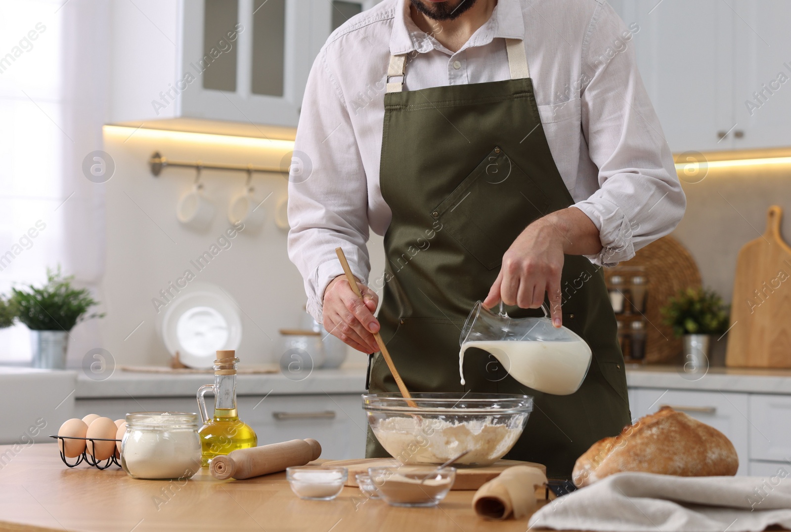 Photo of Making bread. Man pouring milk into bowl with flour at wooden table in kitchen, closeup